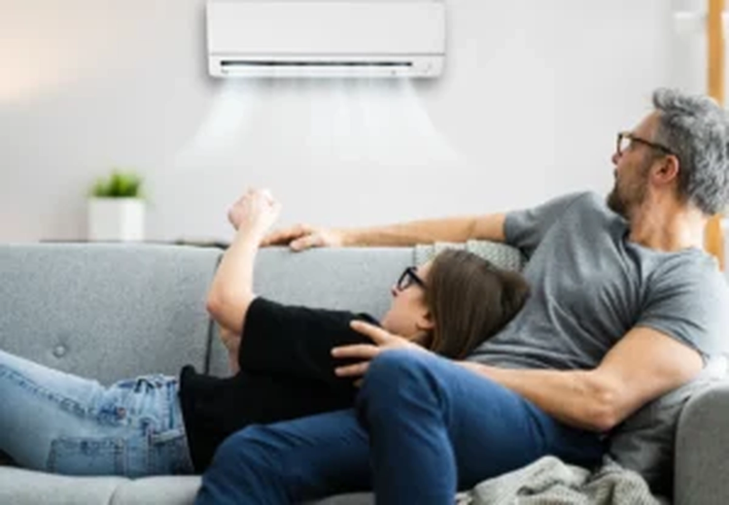 Elektro Wollmer GmbH - Innovative Klimatechnik - Ein Paar sitzt auf dem Sofa vor der an der Wand hängenden Klimaanlage mit Fernbedienung.